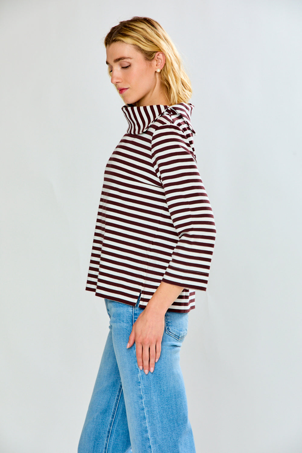 A woman modeling a brown and white horizontally striped knit top with a wide mock neck and three-quarter sleeves, paired with light blue jeans, against a light gray studio background.