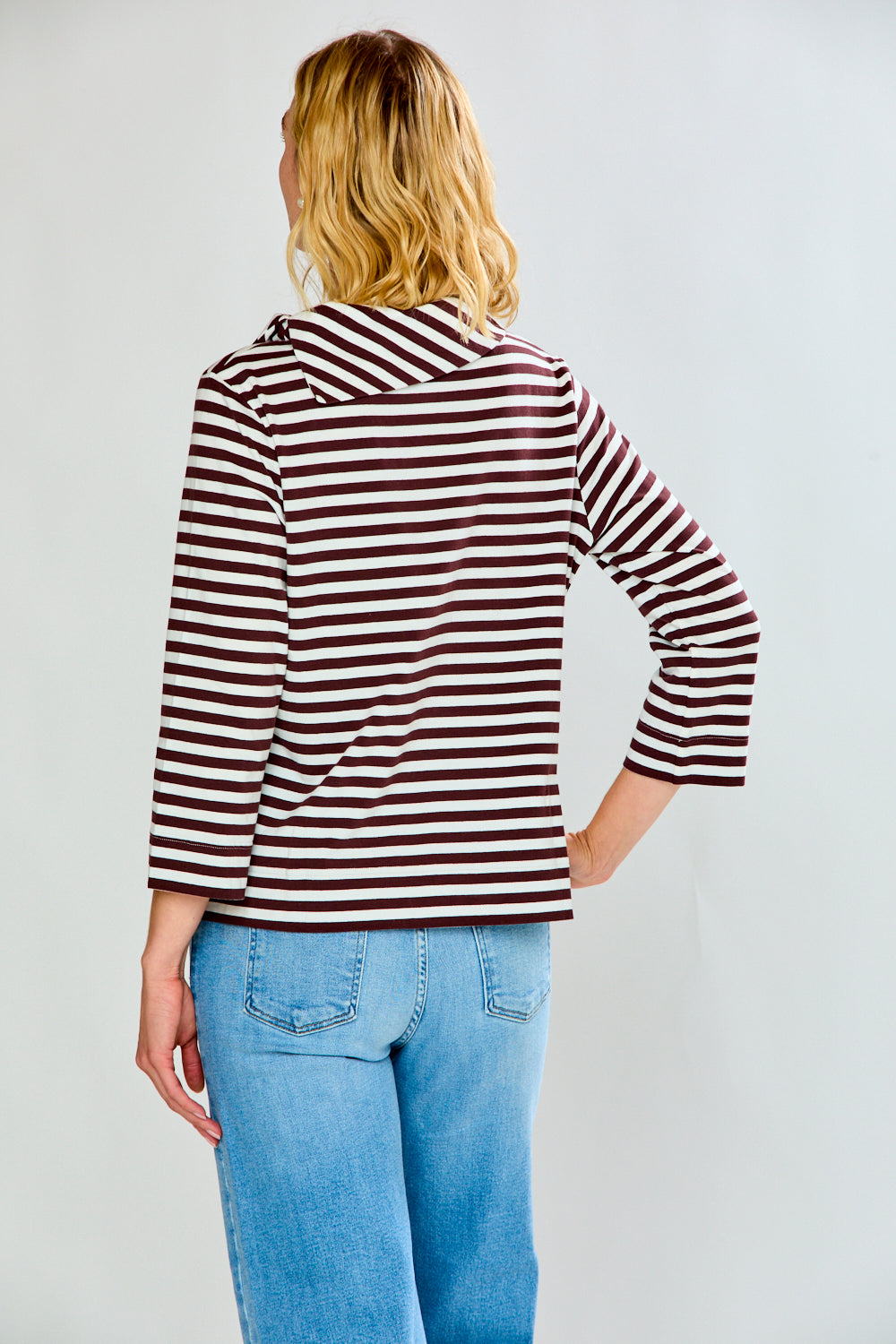 A woman modeling a brown and white horizontally striped knit top with a wide mock neck and three-quarter sleeves, paired with light blue jeans, against a light gray studio background.