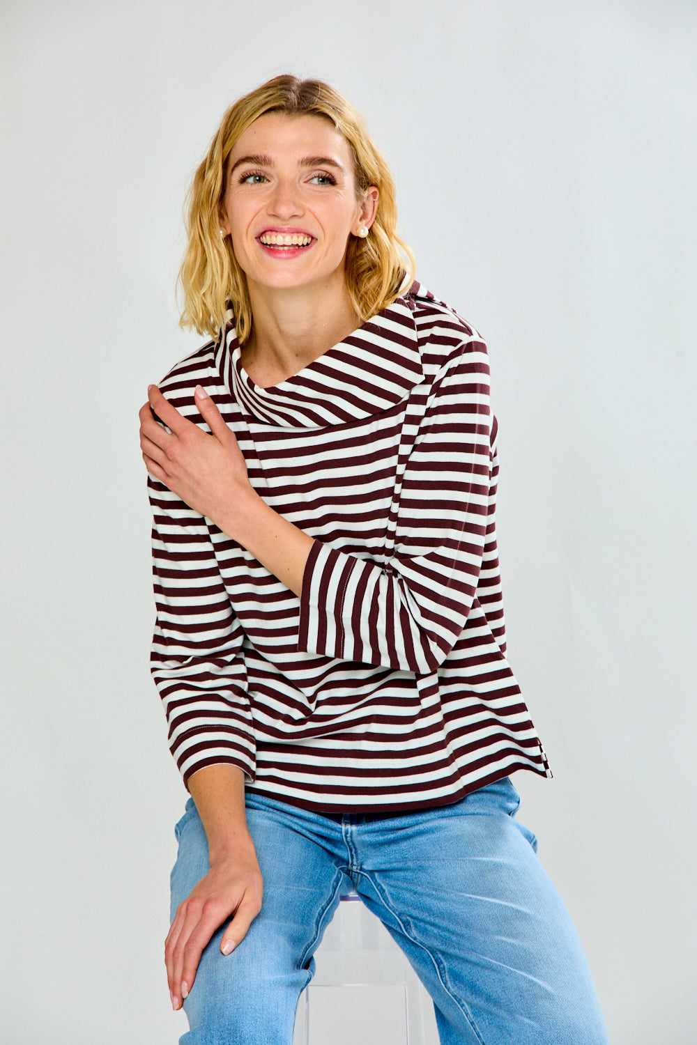A woman modeling a brown and white horizontally striped knit top with a wide mock neck and three-quarter sleeves, paired with light blue jeans, against a light gray studio background.