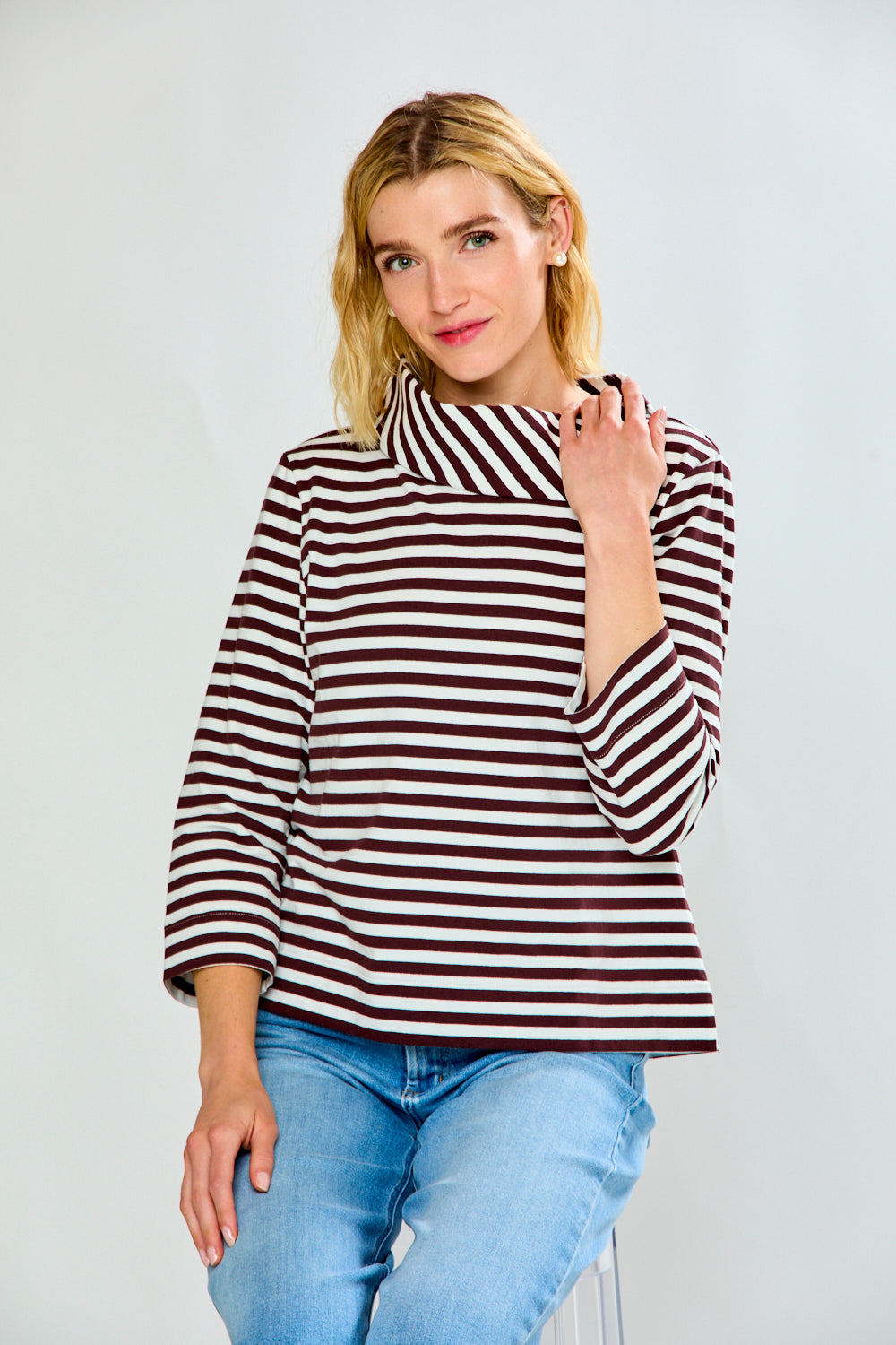A woman modeling a brown and white horizontally striped knit top with a wide mock neck and three-quarter sleeves, paired with light blue jeans, against a light gray studio background.