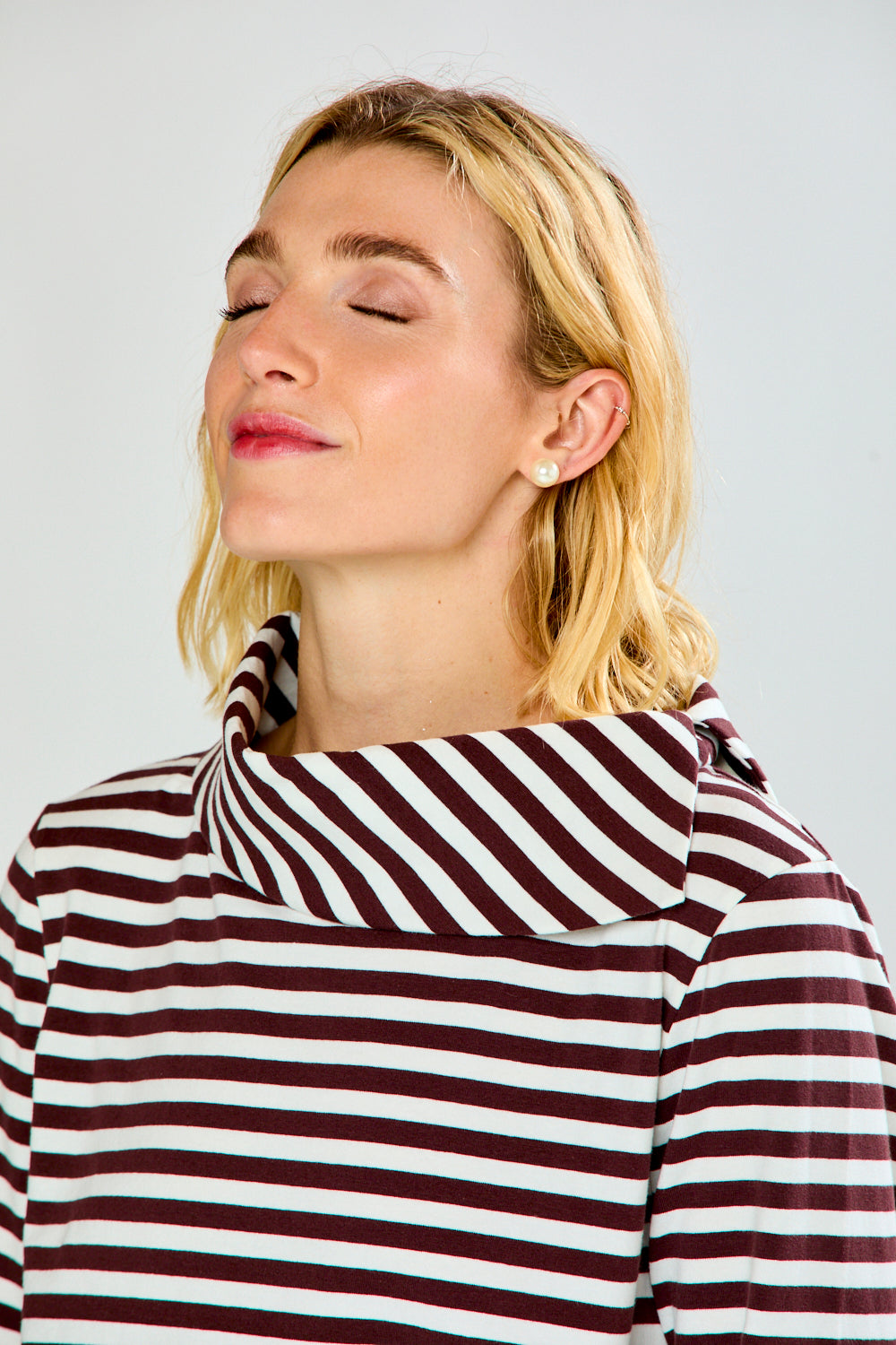 A woman modeling a brown and white horizontally striped knit top with a wide mock neck and three-quarter sleeves, paired with light blue jeans, against a light gray studio background.