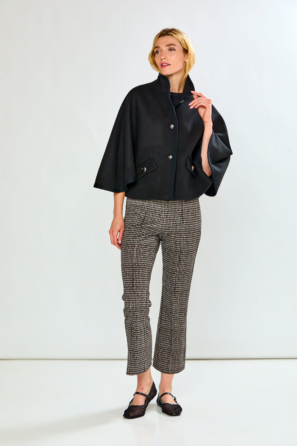A woman modeling a black cape-style jacket with wide sleeves and flap pockets, paired with black-and-white houndstooth pants, against a light gray studio background.