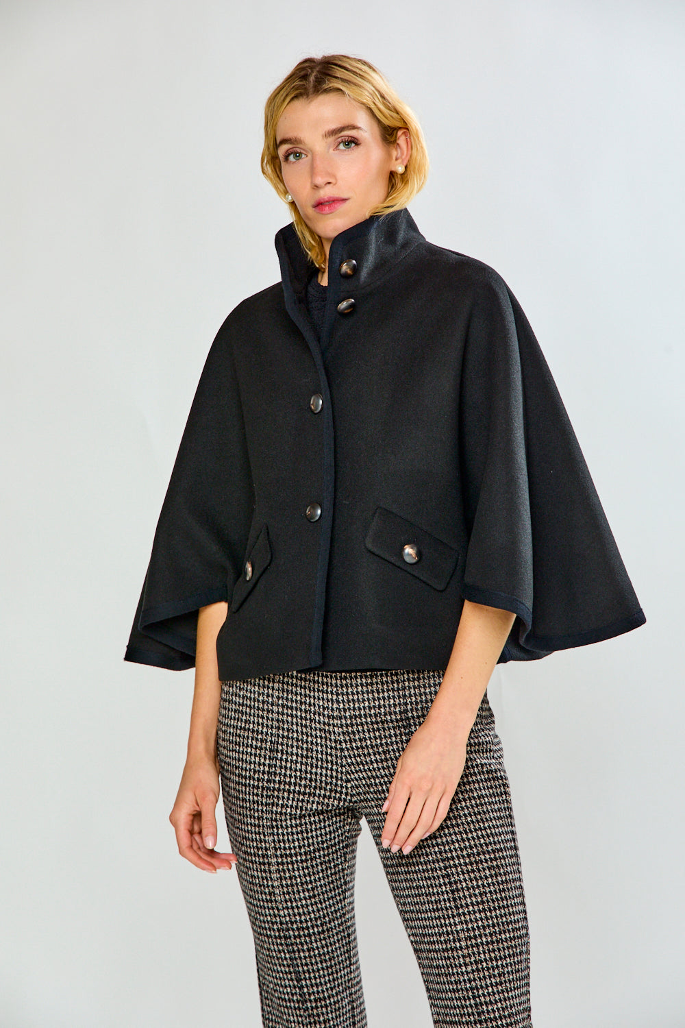 A woman modeling a black cape-style jacket with wide sleeves and flap pockets, paired with black-and-white houndstooth pants, against a light gray studio background.
