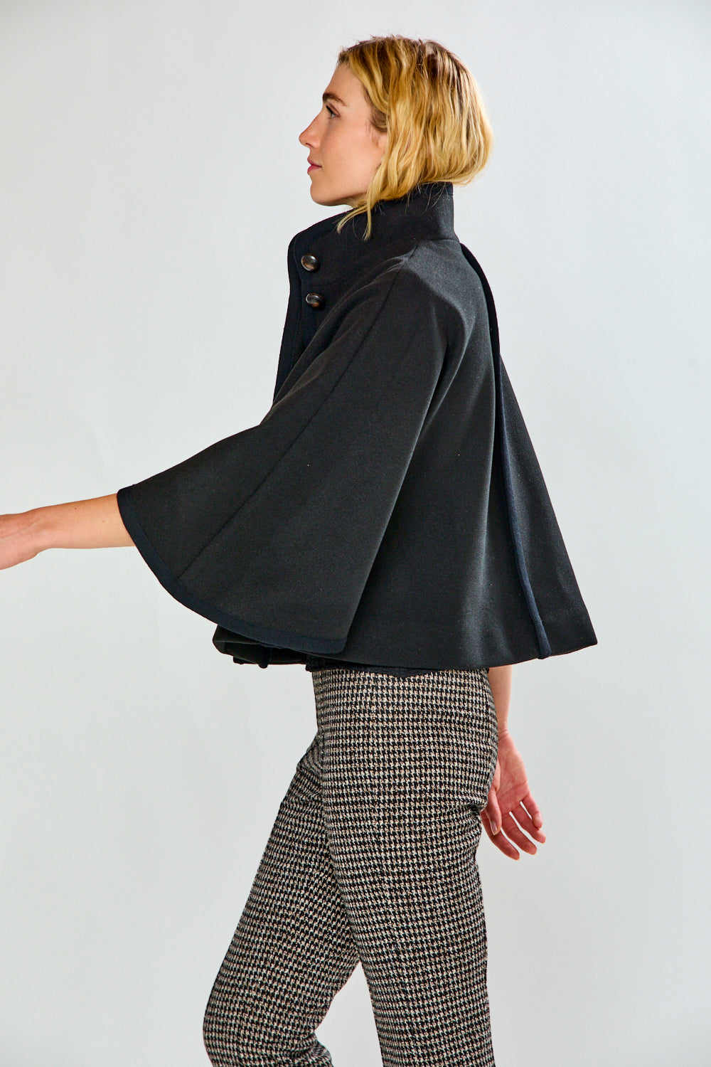 A woman modeling a black cape-style jacket with wide sleeves and flap pockets, paired with black-and-white houndstooth pants, against a light gray studio background.
