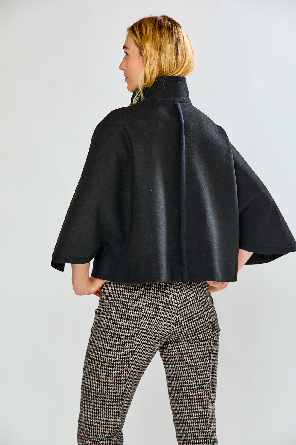 A woman modeling a black cape-style jacket with wide sleeves and flap pockets, paired with black-and-white houndstooth pants, against a light gray studio background.