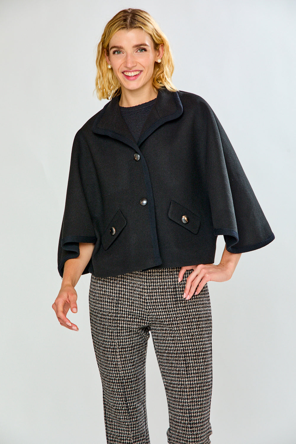 A woman modeling a black cape-style jacket with wide sleeves and flap pockets, paired with black-and-white houndstooth pants, against a light gray studio background.