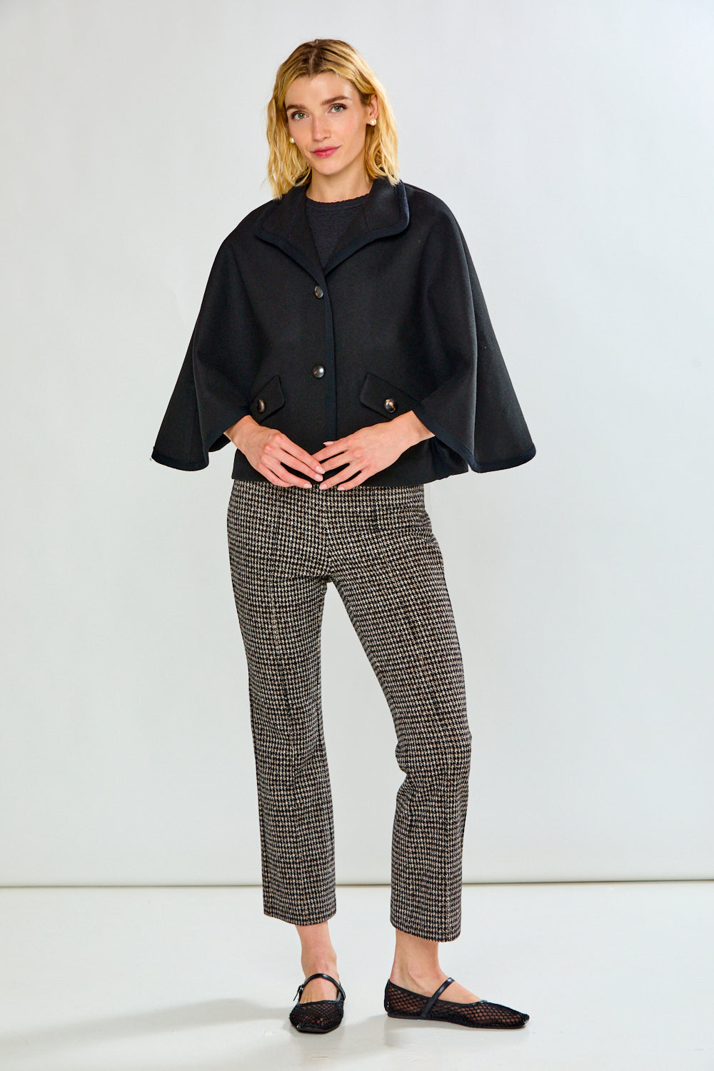 A woman modeling a black cape-style jacket with wide sleeves and flap pockets, paired with black-and-white houndstooth pants, against a light gray studio background.