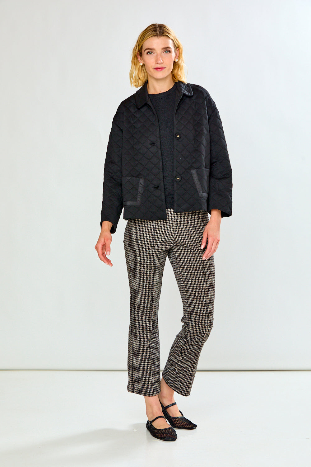A woman modeling a black quilted jacket with front pockets, paired with black and white houndstooth cropped trousers and black flats, against a plain white studio background.