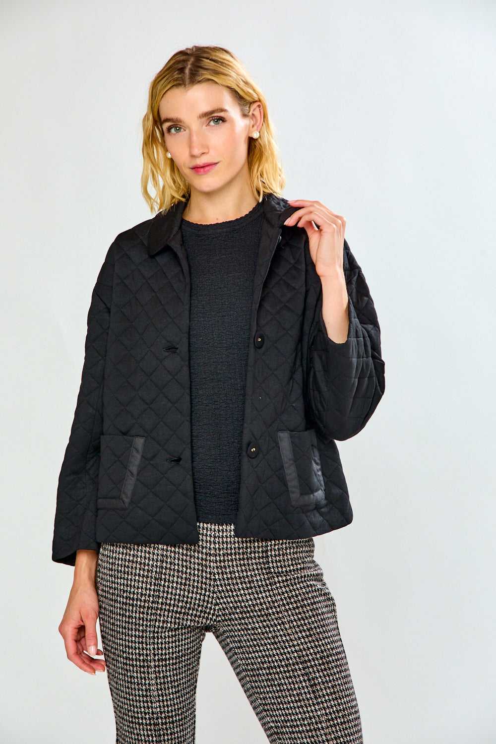 A woman modeling a black quilted jacket with front pockets, paired with black and white houndstooth cropped trousers and black flats, against a plain white studio background.