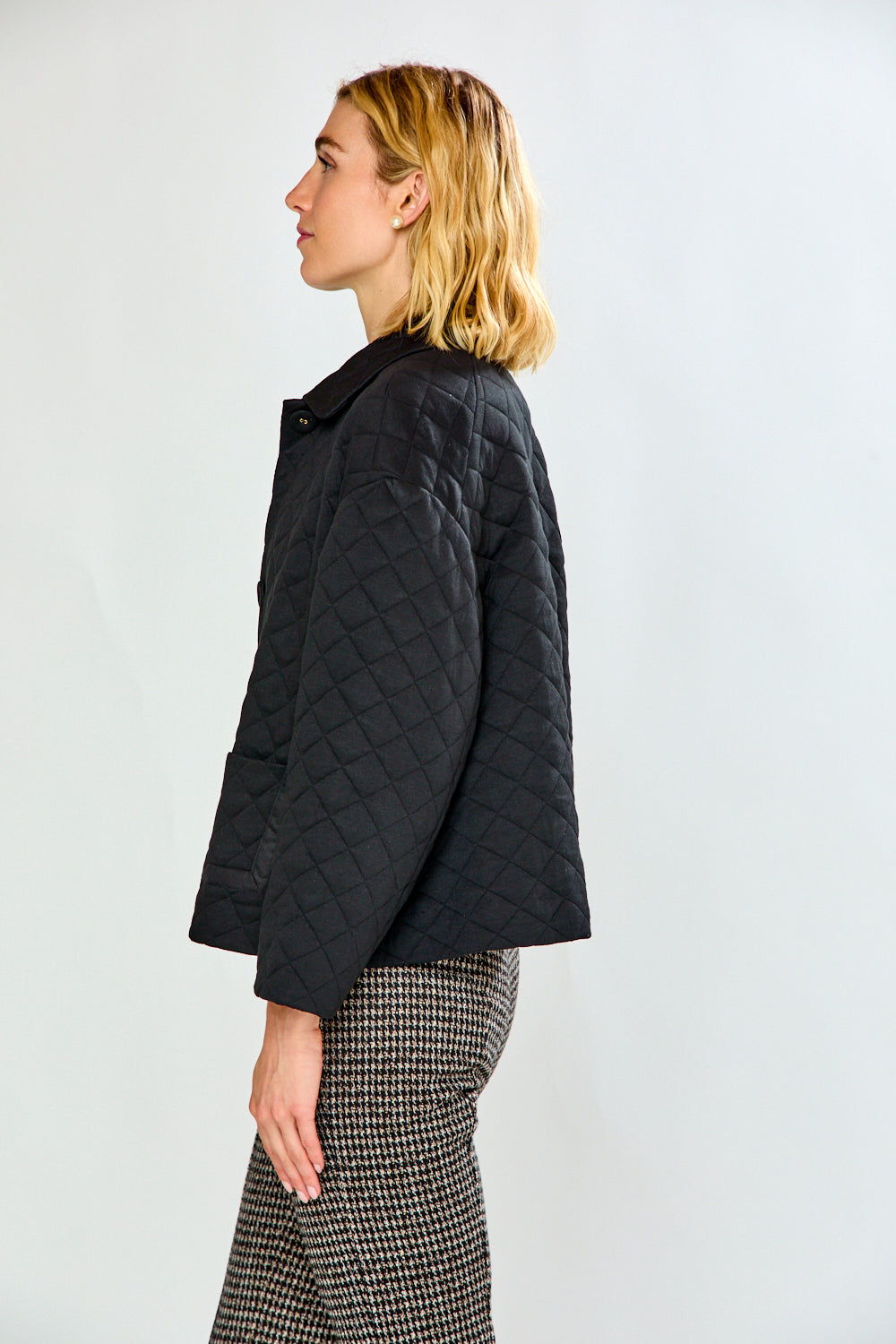 A woman modeling a black quilted jacket with front pockets, paired with black and white houndstooth cropped trousers and black flats, against a plain white studio background.
