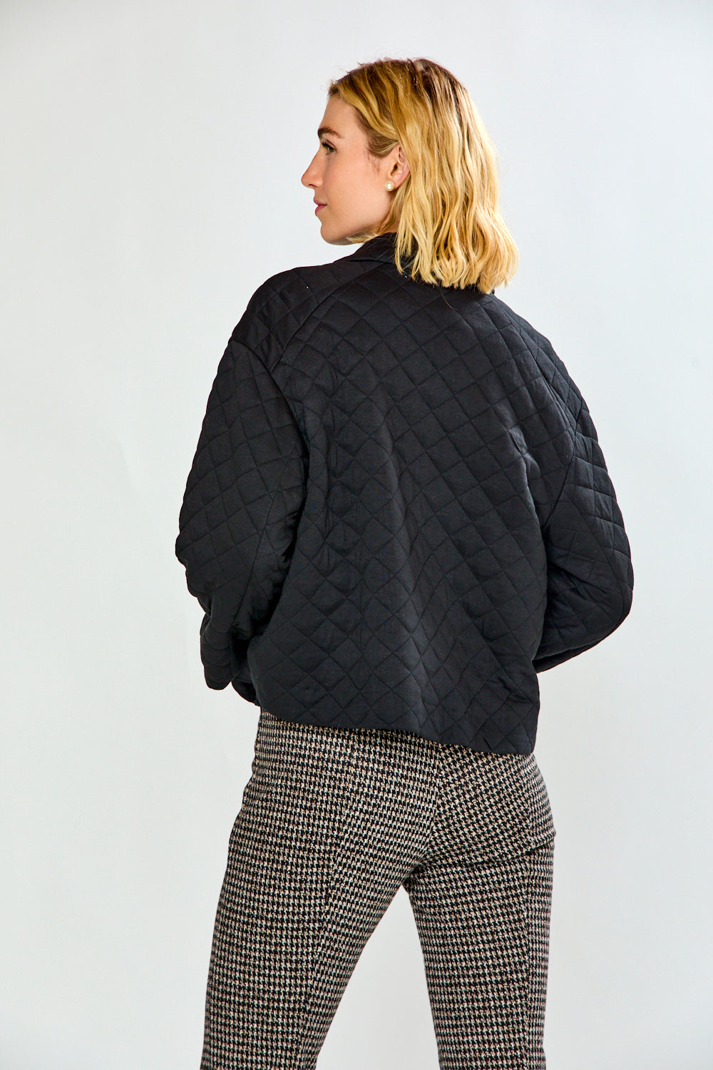 A woman modeling a black quilted jacket with front pockets, paired with black and white houndstooth cropped trousers and black flats, against a plain white studio background.