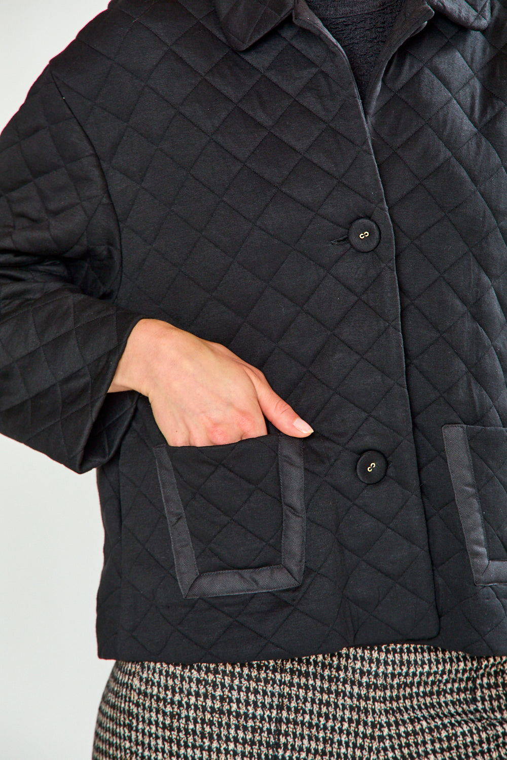 A woman modeling a black quilted jacket with front pockets, paired with black and white houndstooth cropped trousers and black flats, against a plain white studio background.