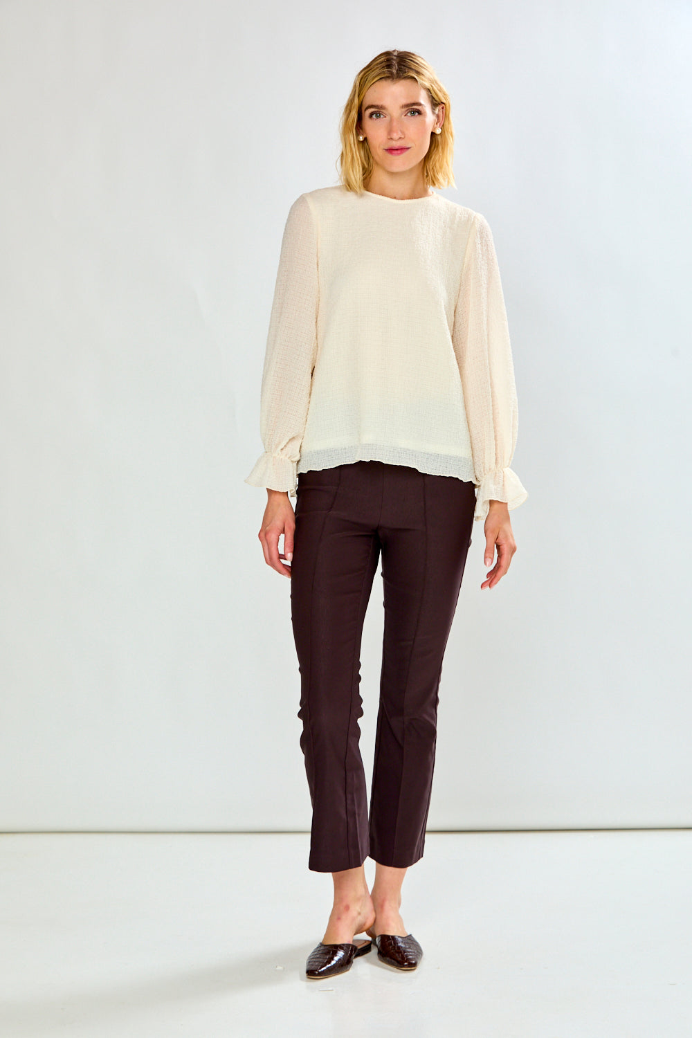 A woman modeling a cream long-sleeve blouse with ruffled cuffs and dark brown cropped trousers, standing against a plain white studio background.