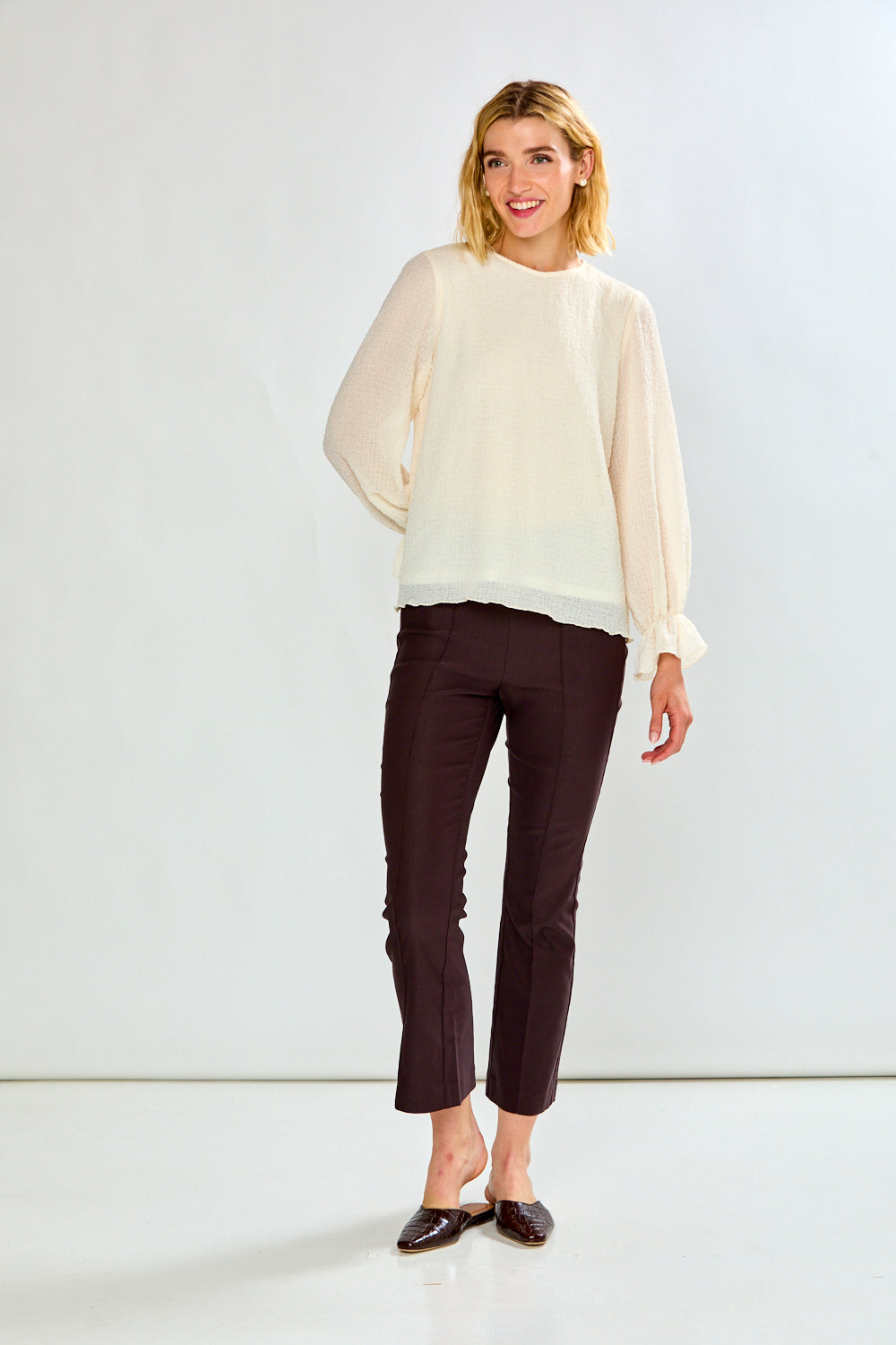 A woman modeling a cream-colored sheer long-sleeve blouse and dark brown cropped trousers, standing against a plain white studio background.
