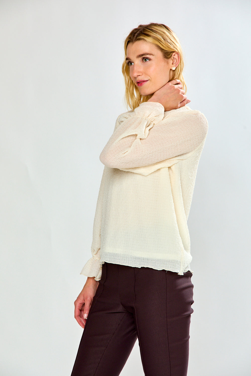 A blonde woman posing in a cream-colored knit blouse  with ruffled sleeves and dark brown pants, set against a plain light gray studio background.