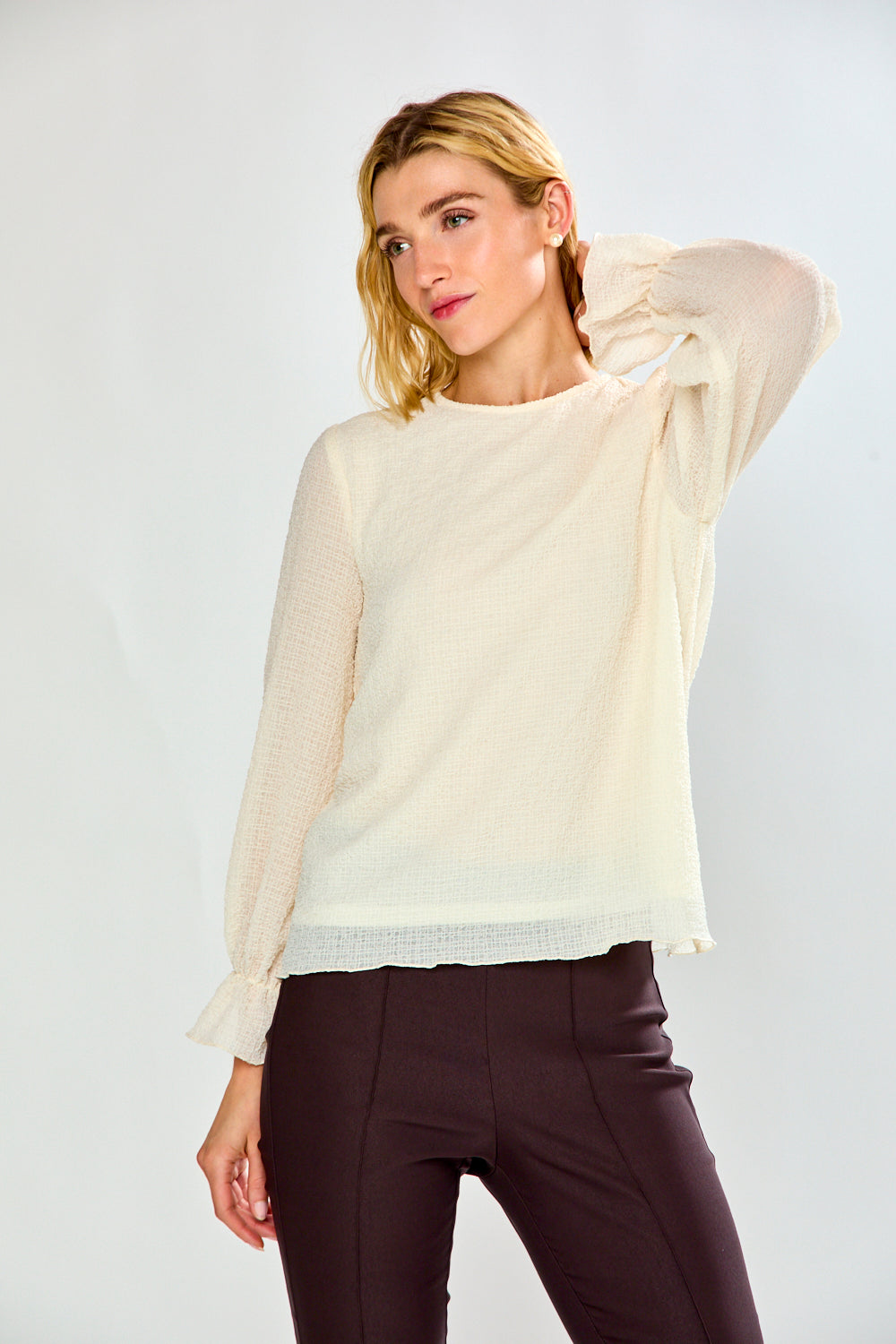 A woman modeling a cream-colored long-sleeve top with  ruffled cuffs, paired with dark brown pants, against a light gray studio background.