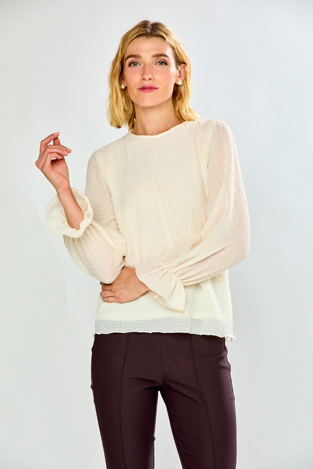 A woman modeling a cream-colored long-sleeve blouse with dramatic gathered sleeves and dark brown trousers, set against a plain light gray studio background.