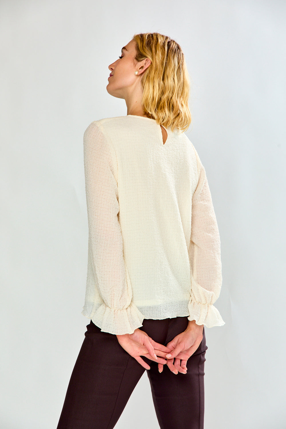 A woman shown from the back wearing a cream-colored textured blouse with long ruffled sleeves and a keyhole back detail, paired with dark brown pants, against a plain light gray background.