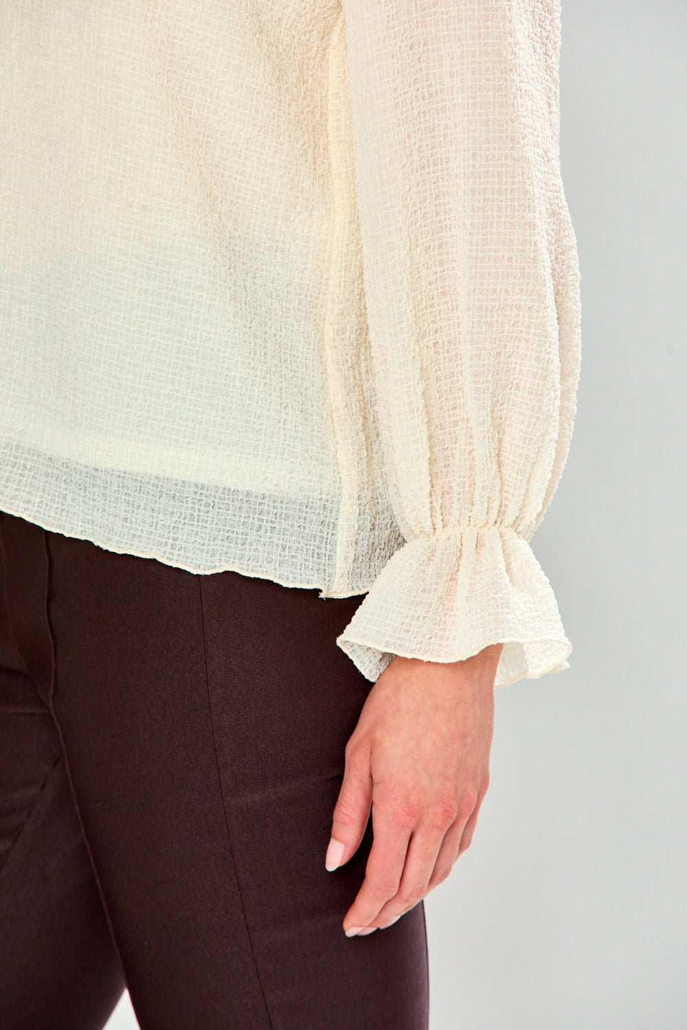 A close-up of a person wearing a cream-colored, semi-sheer long-sleeve blouse with a gathered ruffled cuff, paired with dark brown pants, against a light gray background.