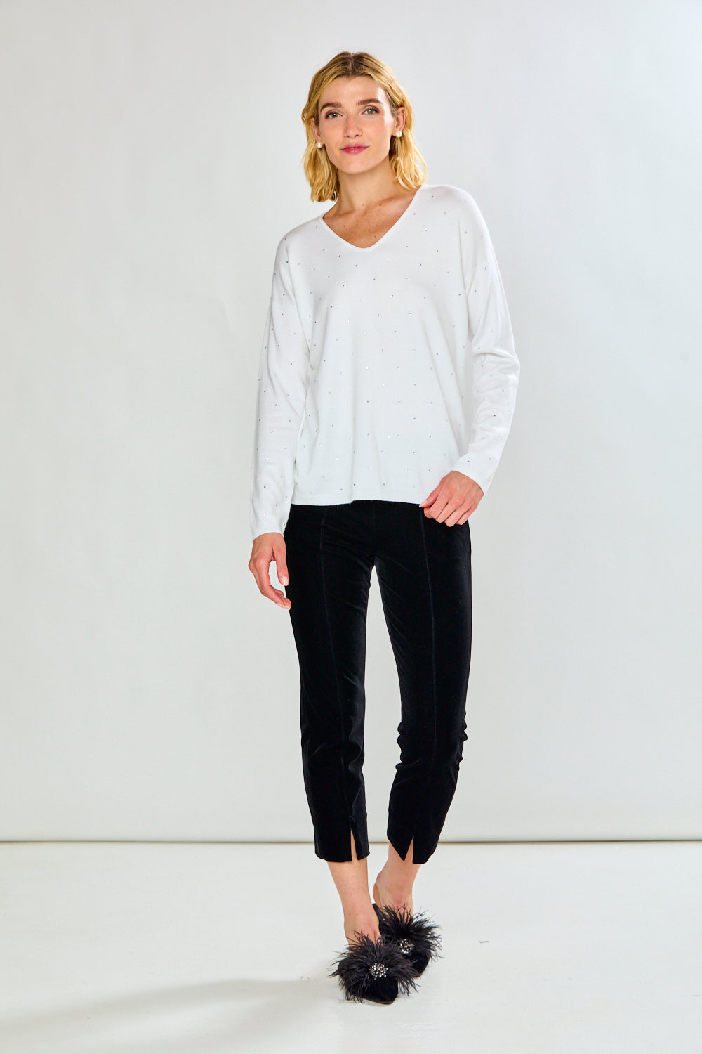 A woman modeling a white long-sleeve V-neck knit top with tiny crystal details, paired with black pants, against a light gray studio background.