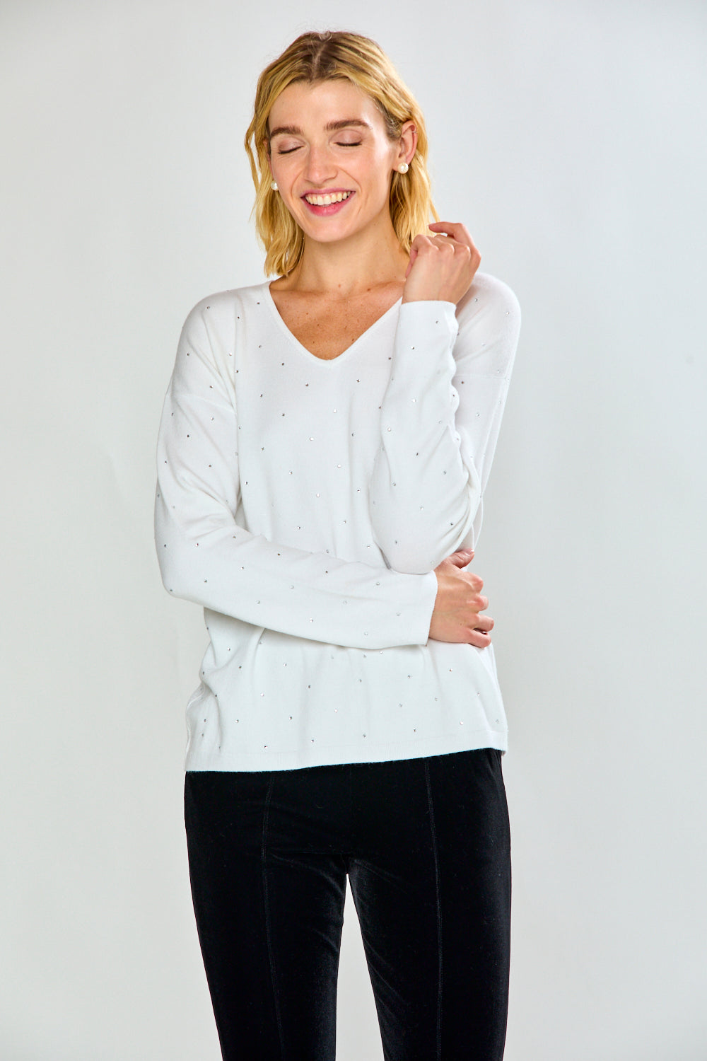 A woman modeling a white long-sleeve V-neck knit top with tiny crystal details, paired with black pants, against a light gray studio background.