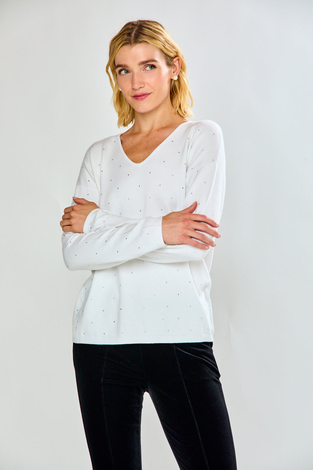 A woman modeling a white long-sleeve V-neck knit top with tiny crystal details, paired with black pants, against a light gray studio background.