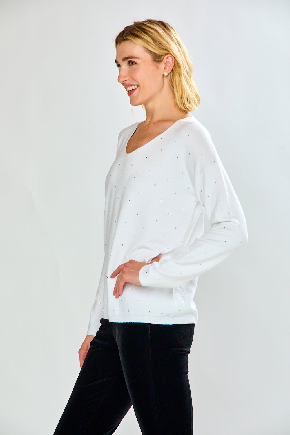 A woman modeling a white long-sleeve V-neck knit top with tiny crystal details, paired with black pants, against a light gray studio background.