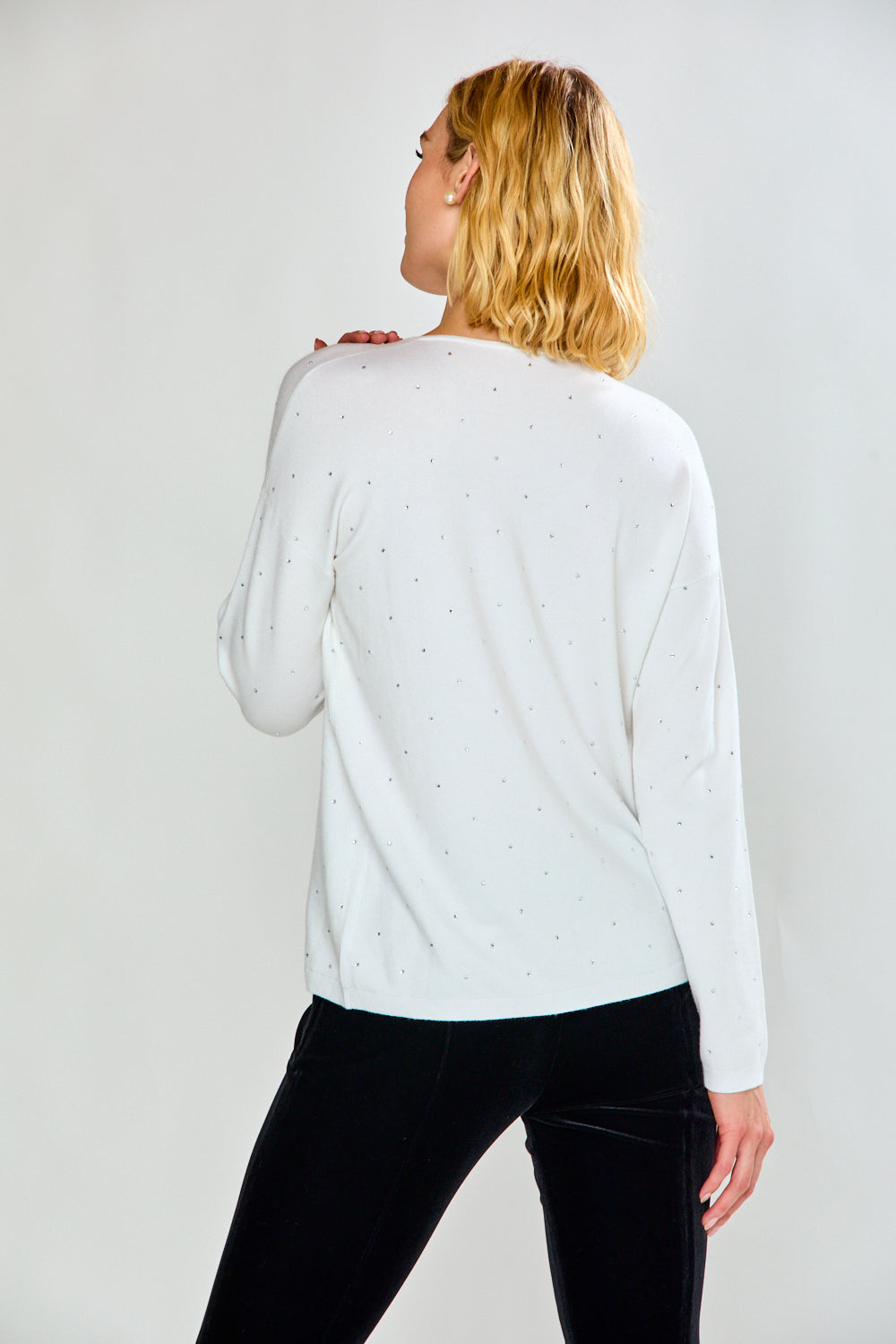 A woman modeling a white long-sleeve V-neck knit top with tiny crystal details, paired with black pants, against a light gray studio background.