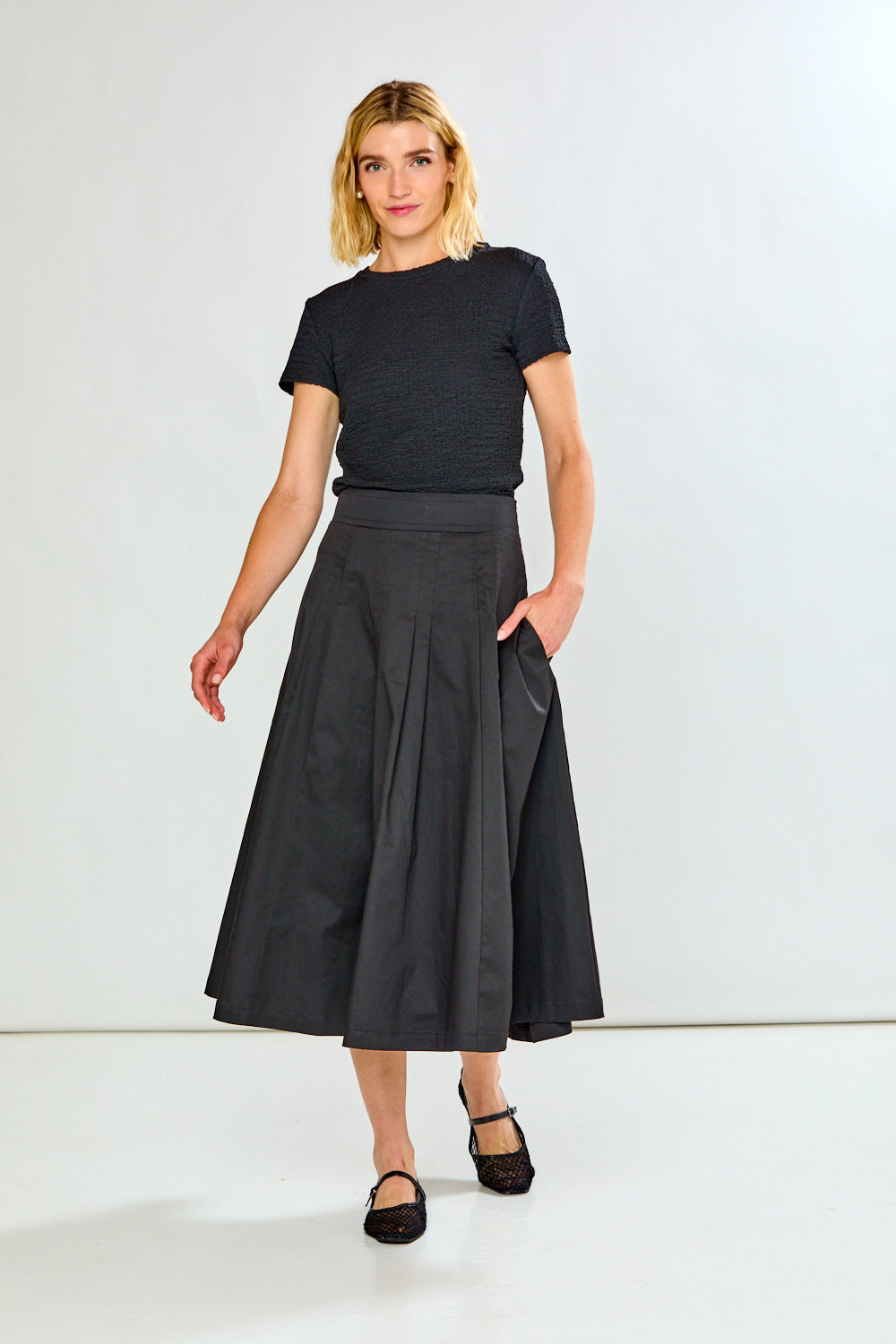 A woman modeling a black short-sleeve textured top and a high-waisted, pleated black midi skirt with side pockets, paired with black flats, against a plain white studio background.