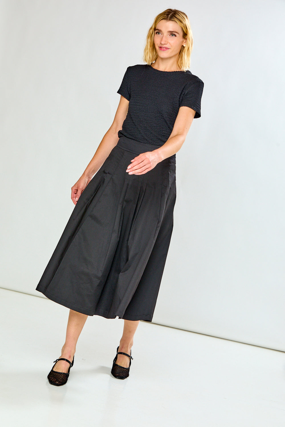 A woman modeling a black short-sleeve textured top and a high-waisted, pleated black midi skirt with side pockets, paired with black flats, against a plain white studio background.