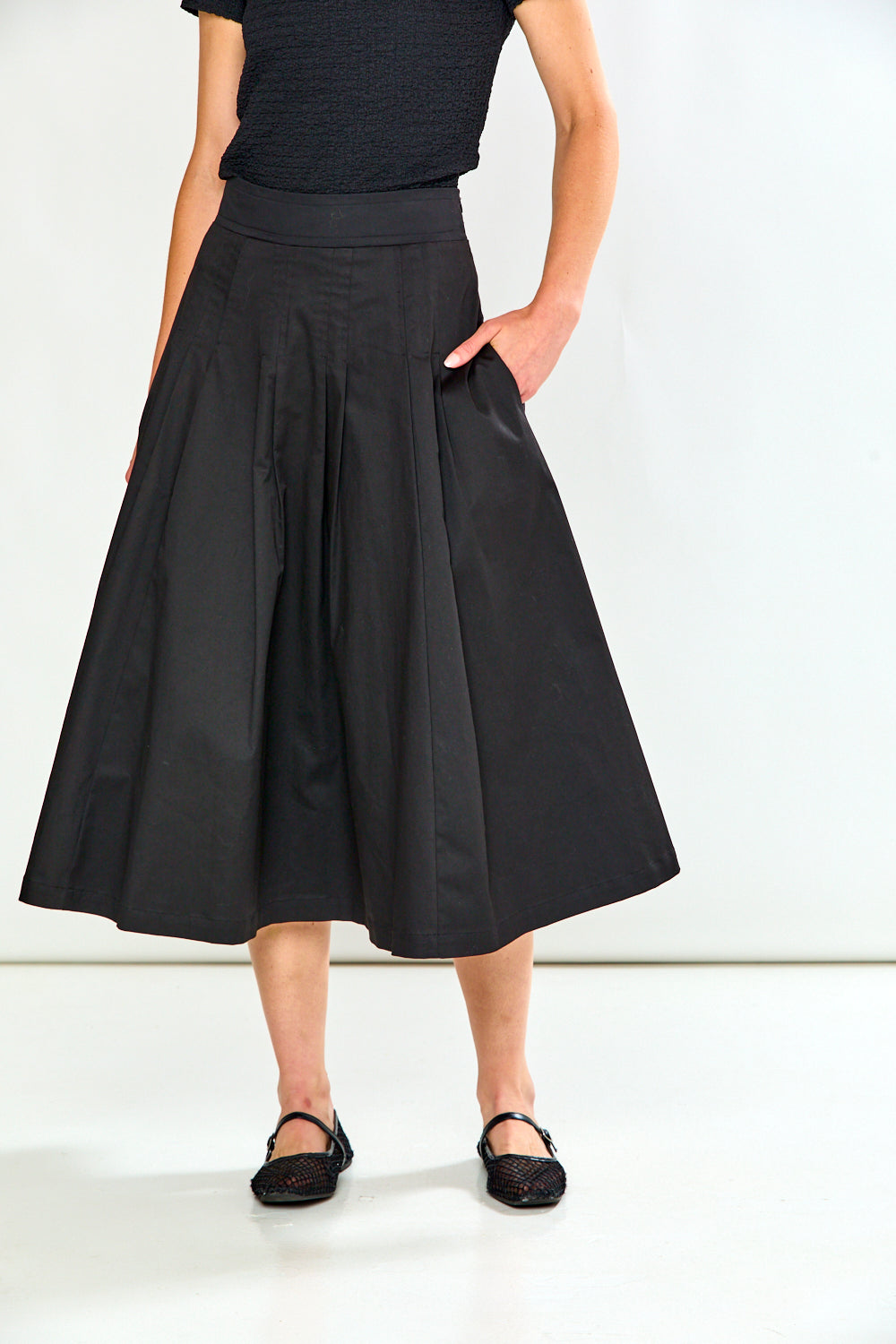A model wearing a black high-waisted, pleated A-line midi skirt with side pockets, paired with a black top and black flat shoes, against a plain white studio background.