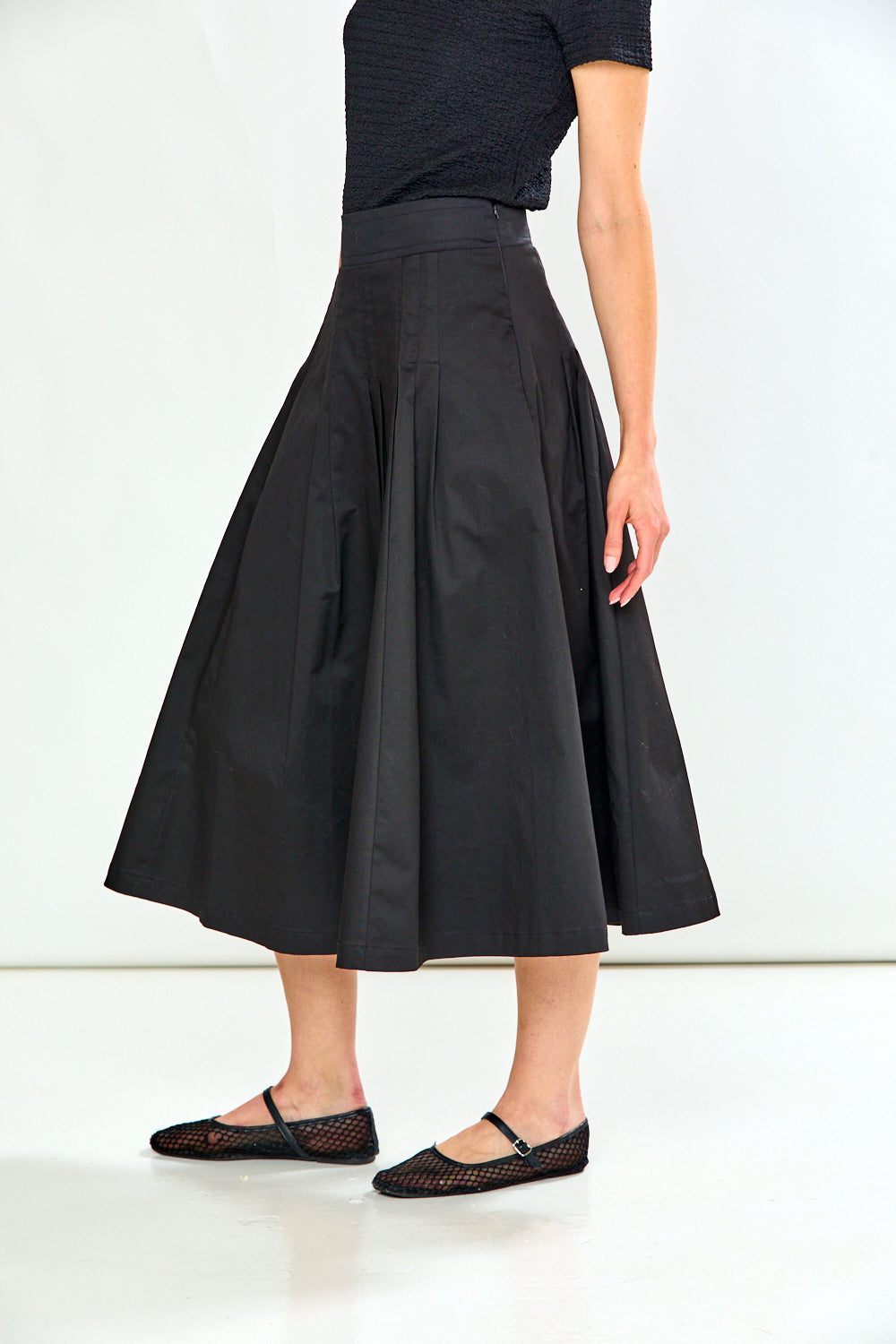 A model wearing a black high-waisted, flared midi skirt paired with a black short-sleeve top and black flat shoes, shown from the waist down against a white studio background.