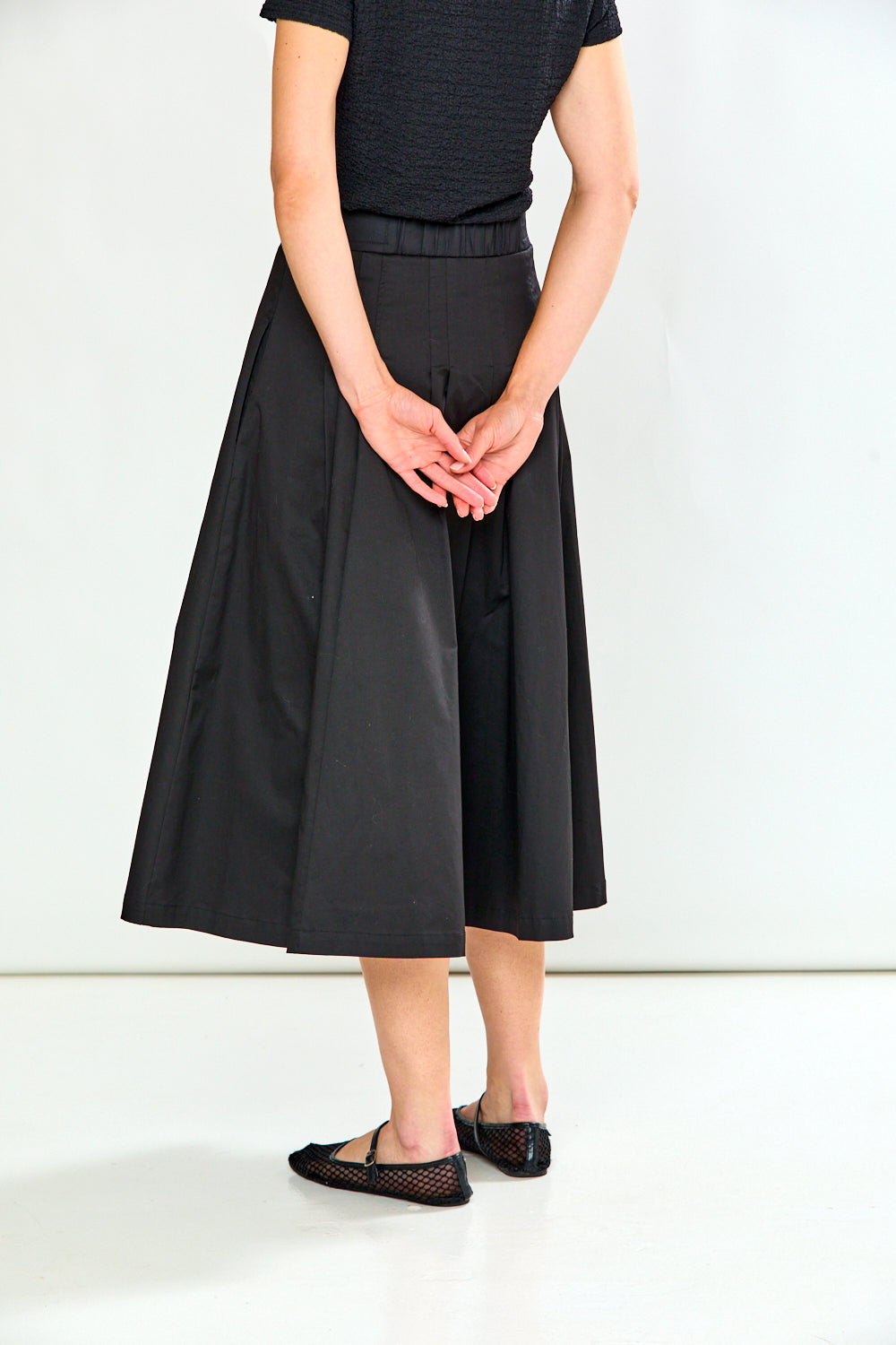 A person shown from the back wearing a black midi skirt with a gathered waist, paired with a black ribbed short-sleeve top and black flats, against a plain white studio background.