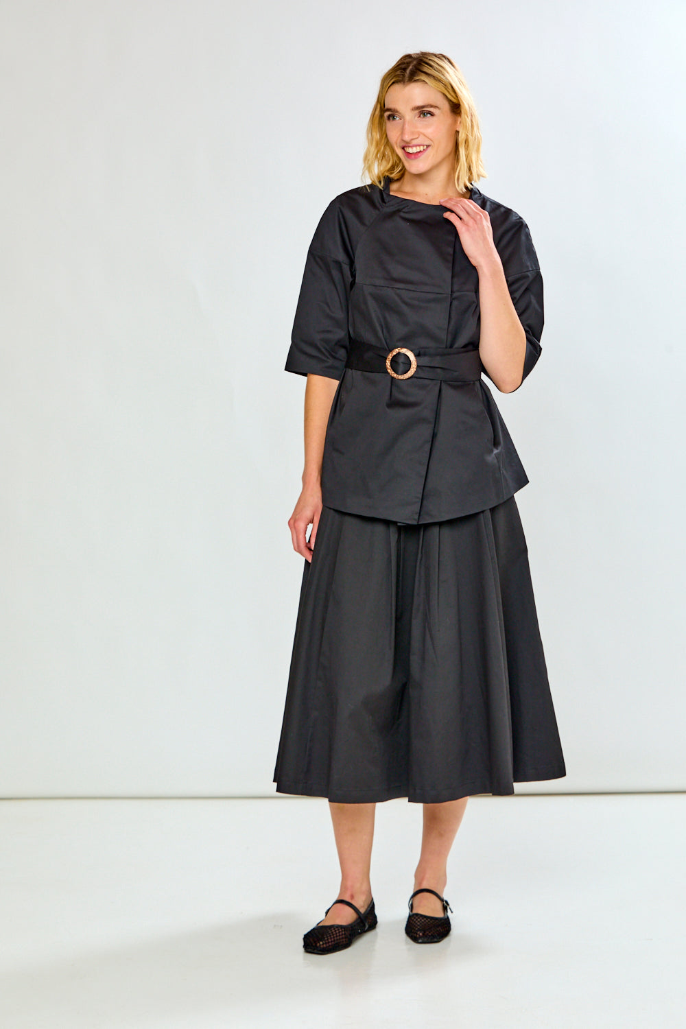 A woman modeling a black short-sleeve textured top and a high-waisted, pleated black midi skirt with side pockets, paired with black flats, against a plain white studio background.