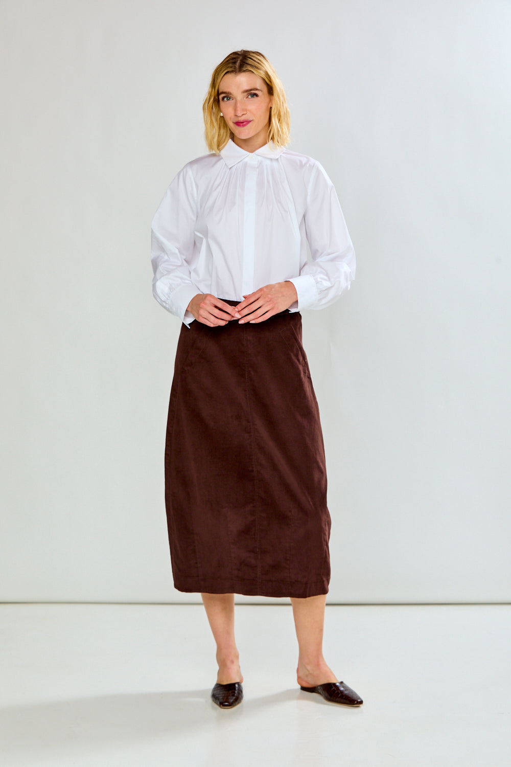 A model wearing a dark brown corduroy midi skirt with a white long-sleeve shirt and dark brown pointed-toe flats, standing against a plain white studio background.