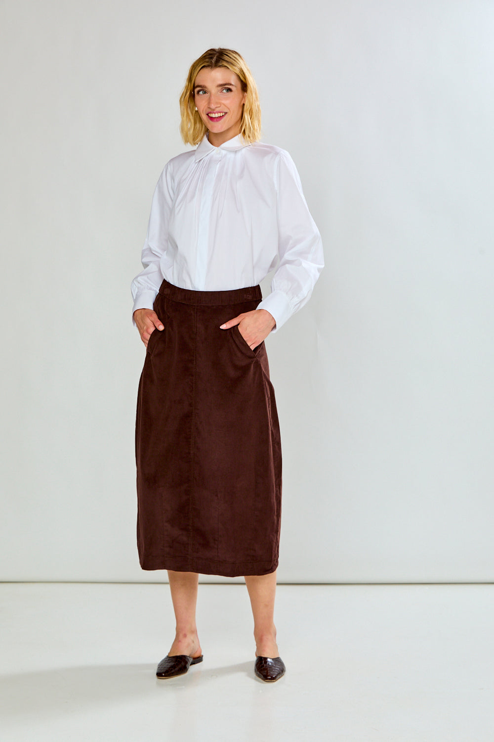 A woman modeling a white long-sleeve blouse and a dark brown midi skirt with front patch pockets, standing against a plain light gray studio background.