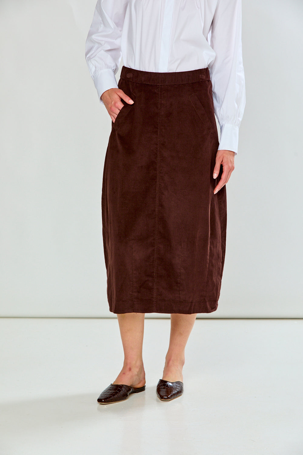 A model wearing a dark brown corduroy midi skirt with a white long-sleeve shirt and dark brown pointed-toe flats, standing against a plain white studio background.