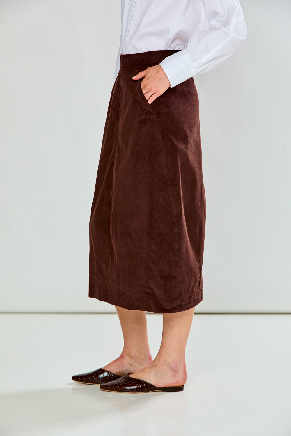 A model wearing a dark brown corduroy midi skirt with a white long-sleeve shirt and dark brown pointed-toe flats, standing against a plain white studio background.