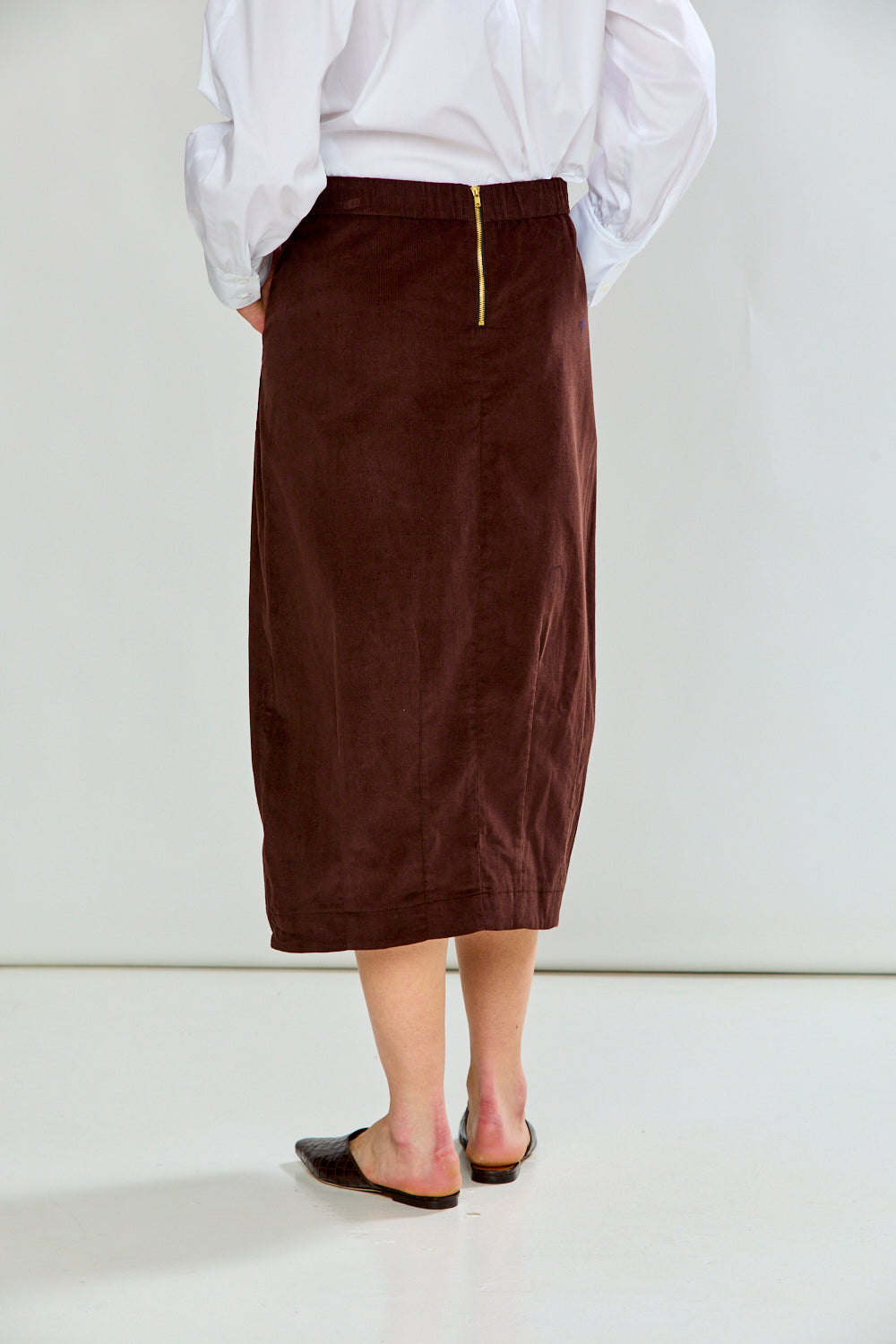 A person shown from the back wearing a dark brown corduroy midi skirt with a gold zipper at the waist, paired with a white long-sleeve blouse and dark flat shoes, against a plain white studio background.