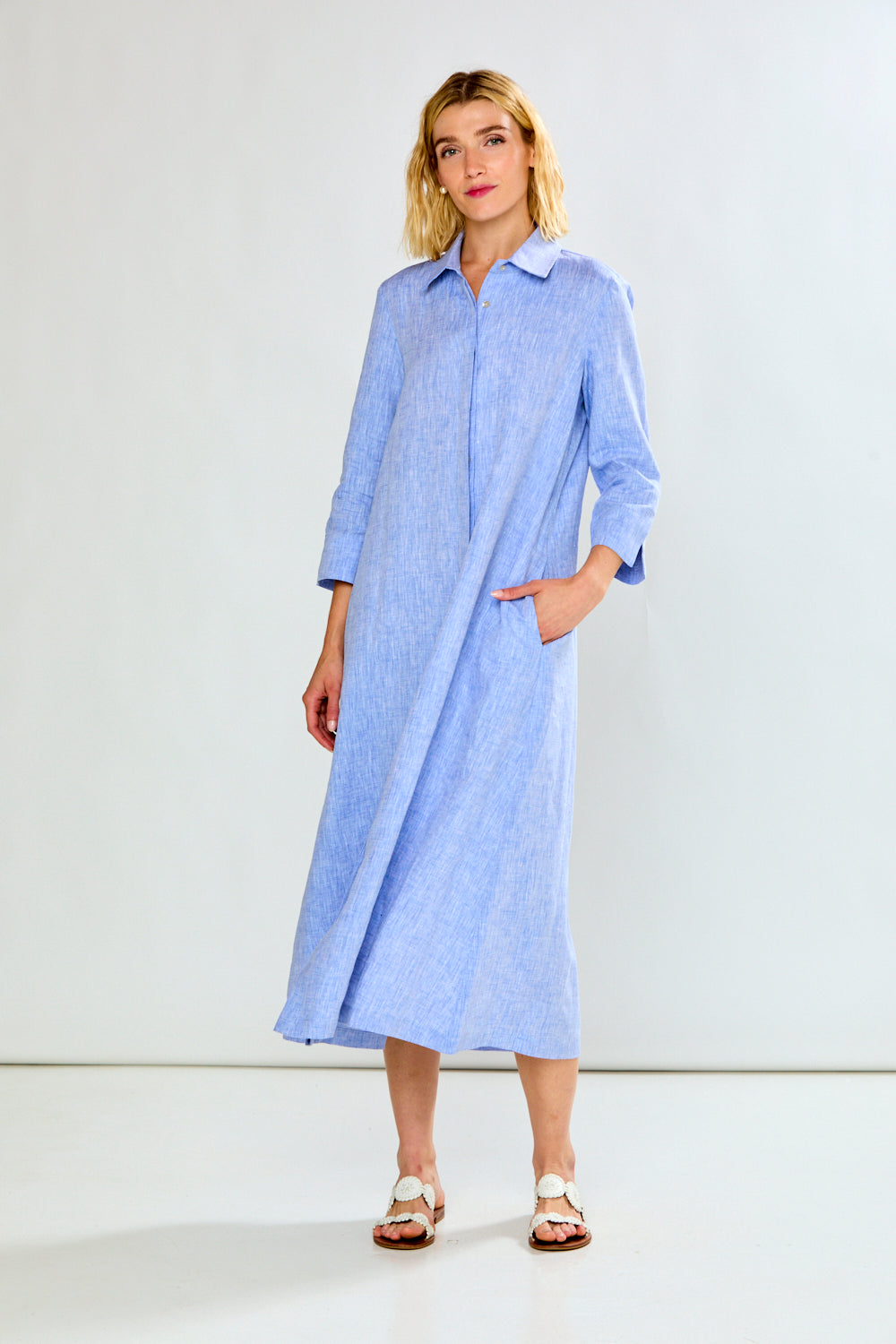 A woman modeling a light blue, long-sleeved midi shirt dress with a collar and front pockets, standing against a plain white studio background.