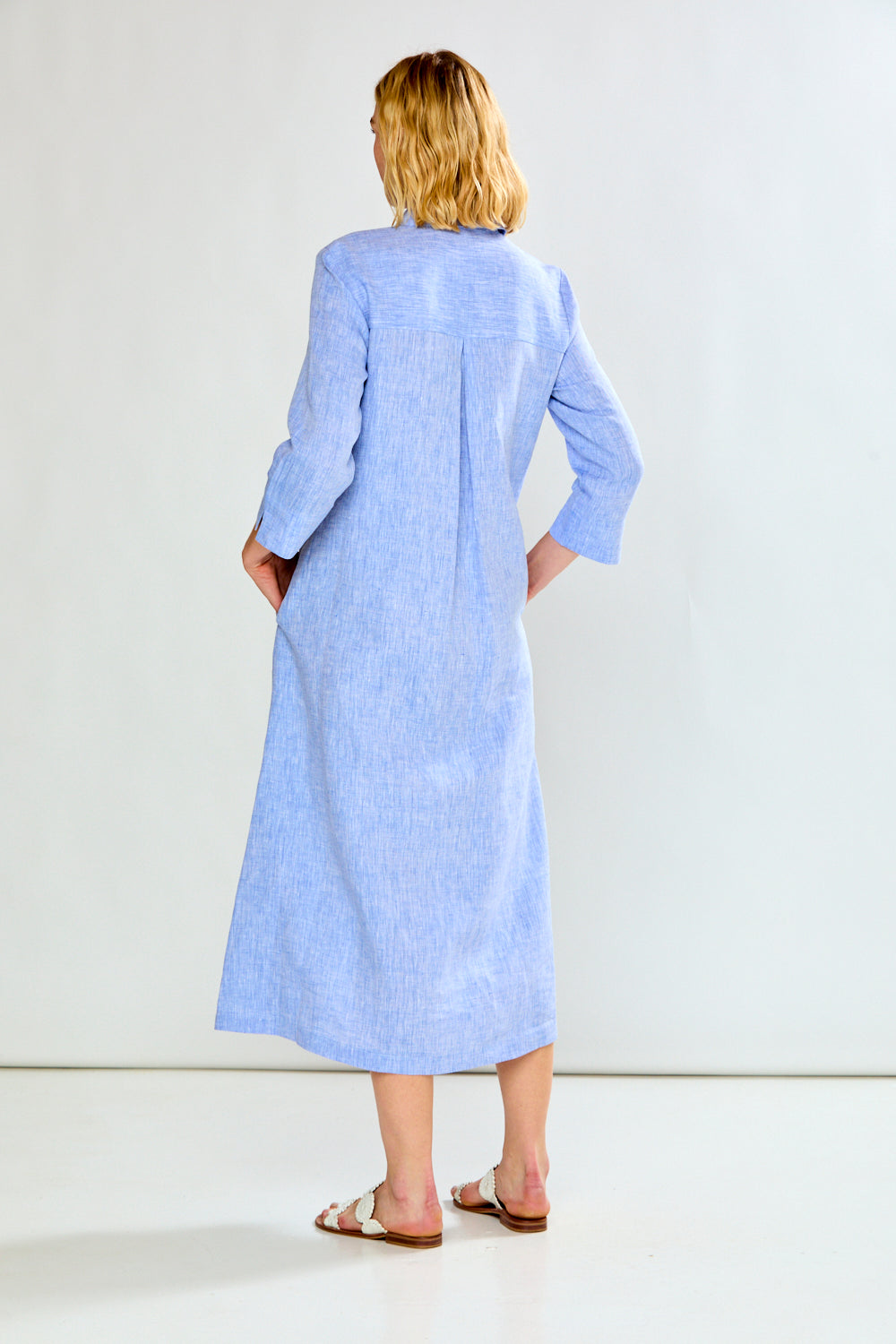 A woman viewed from behind wearing a light blue long-sleeve linen midi dress with pockets, paired with brown leather slide sandals, set against a plain white studio background.