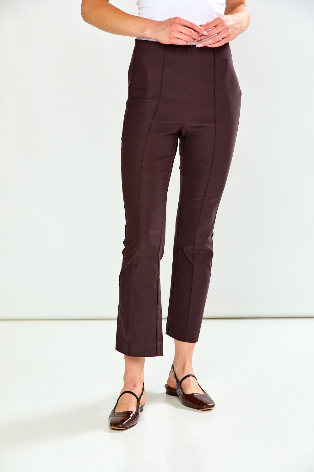 A person shown from the waist down wearing dark brown cropped flare pants and dark flat sandals against a clean white studio background.