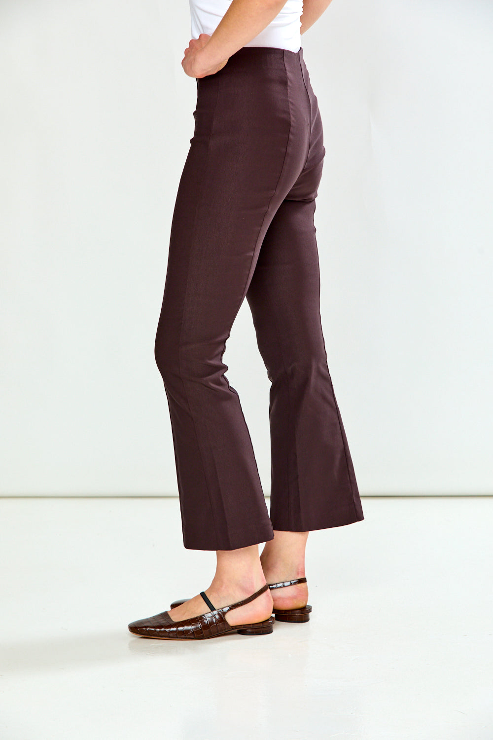 A person shown from the waist down wearing dark brown cropped flare pants and dark flat sandals against a clean white studio background.