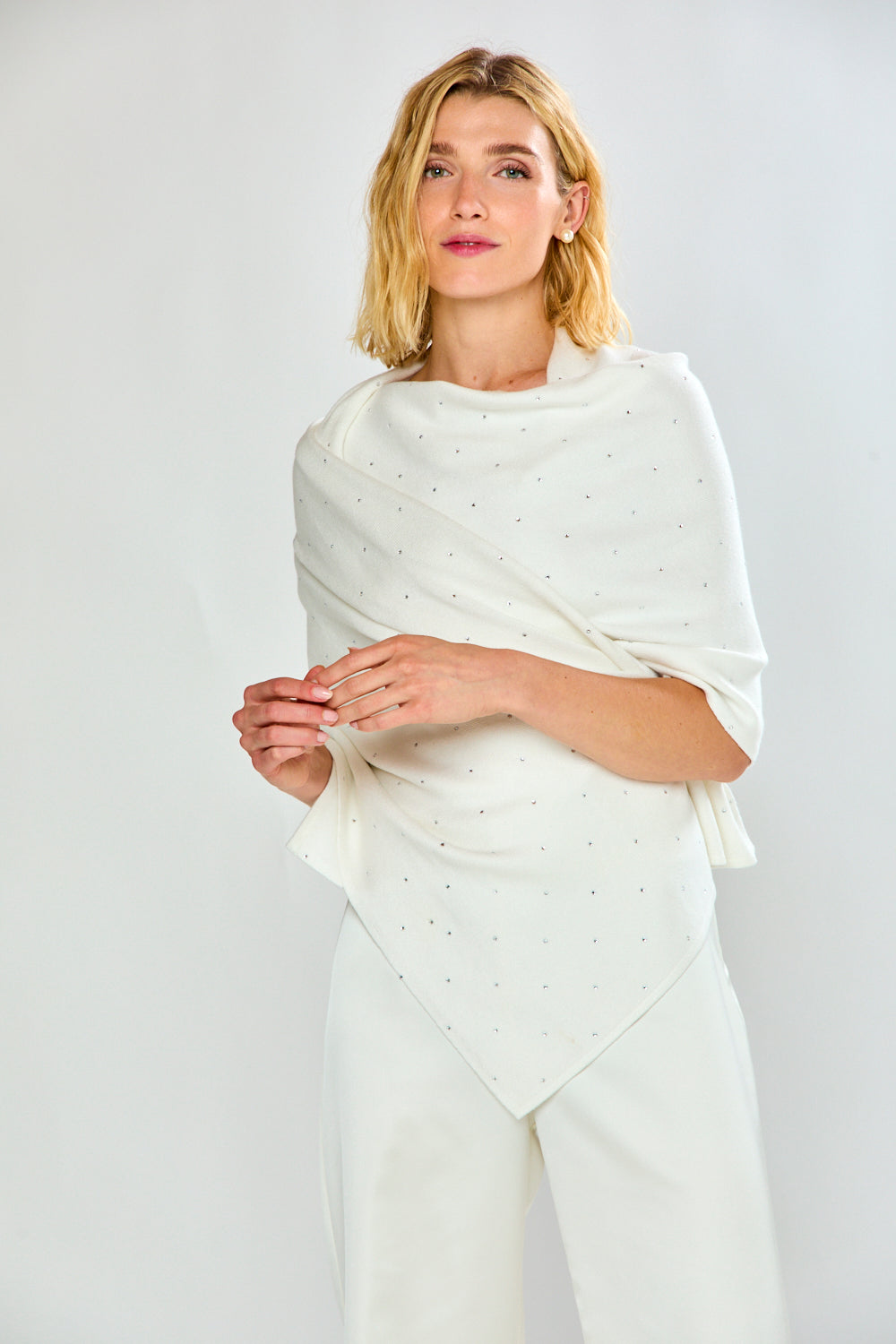 A woman modeling a white knit shawl with tiny silver rhinestone accents, worn over a white top and white trousers, against a light gray studio background.
