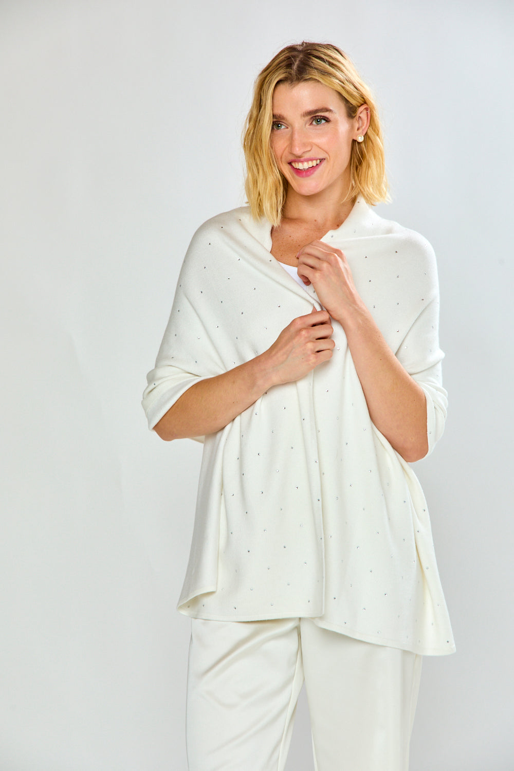 A woman modeling a white knit shawl with tiny silver rhinestone accents, worn over a white top and white trousers, against a light gray studio background.