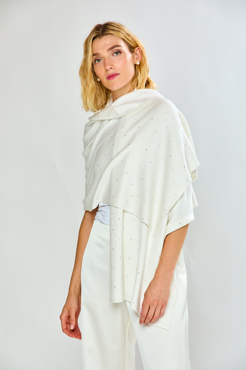 A woman modeling a white knit shawl with tiny silver rhinestone accents, worn over a white top and white trousers, against a light gray studio background.