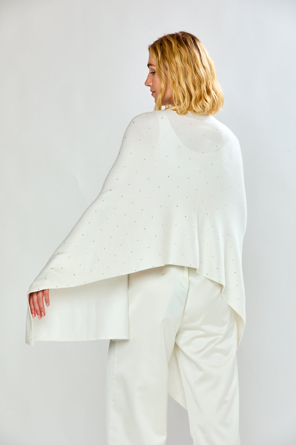 A woman modeling a white knit shawl with tiny silver rhinestone accents, worn over a white top and white trousers, against a light gray studio background.