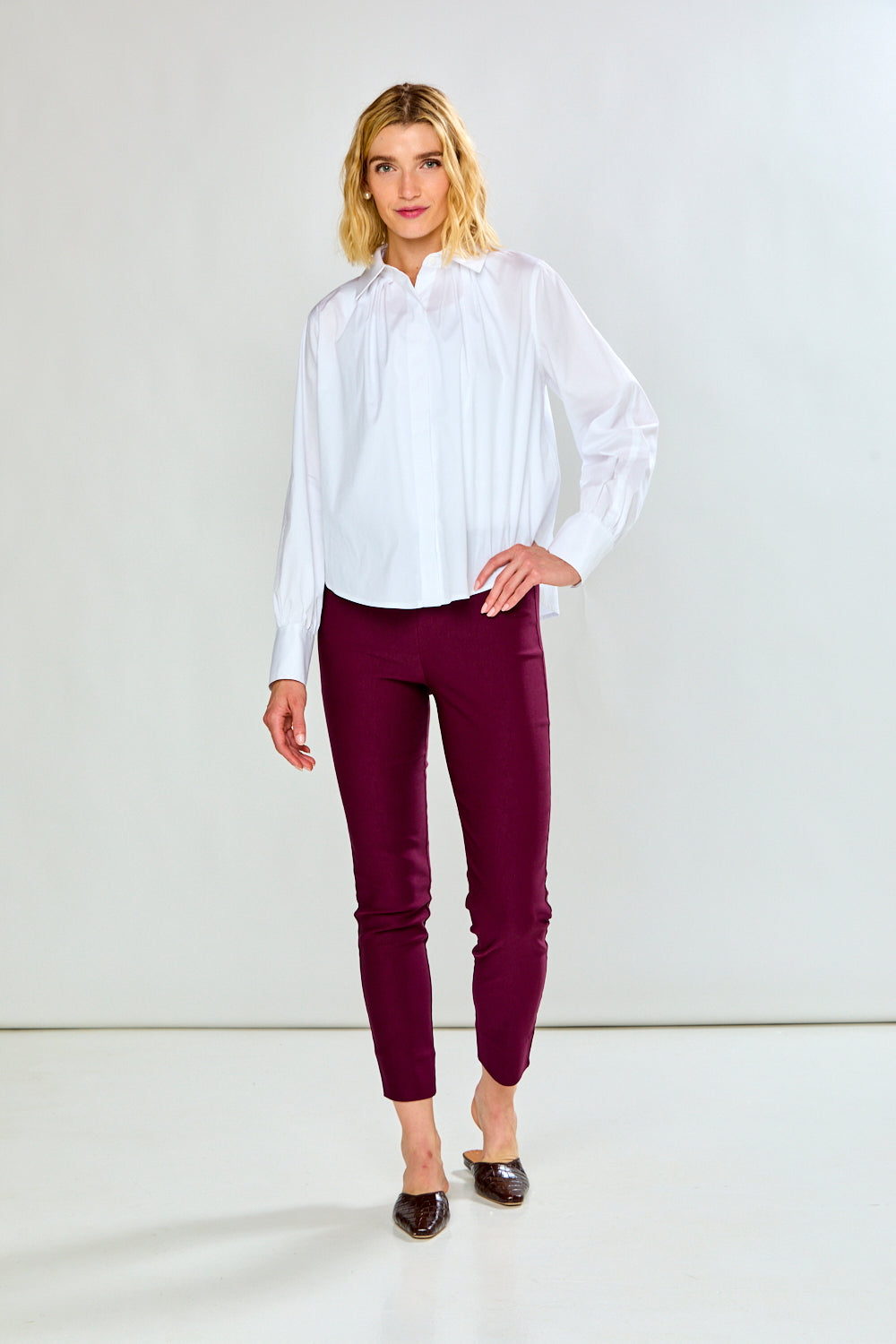 A woman modeling a white long-sleeve button-up blouse and dark burgundy cropped pants, standing against a plain light gray studio background.
