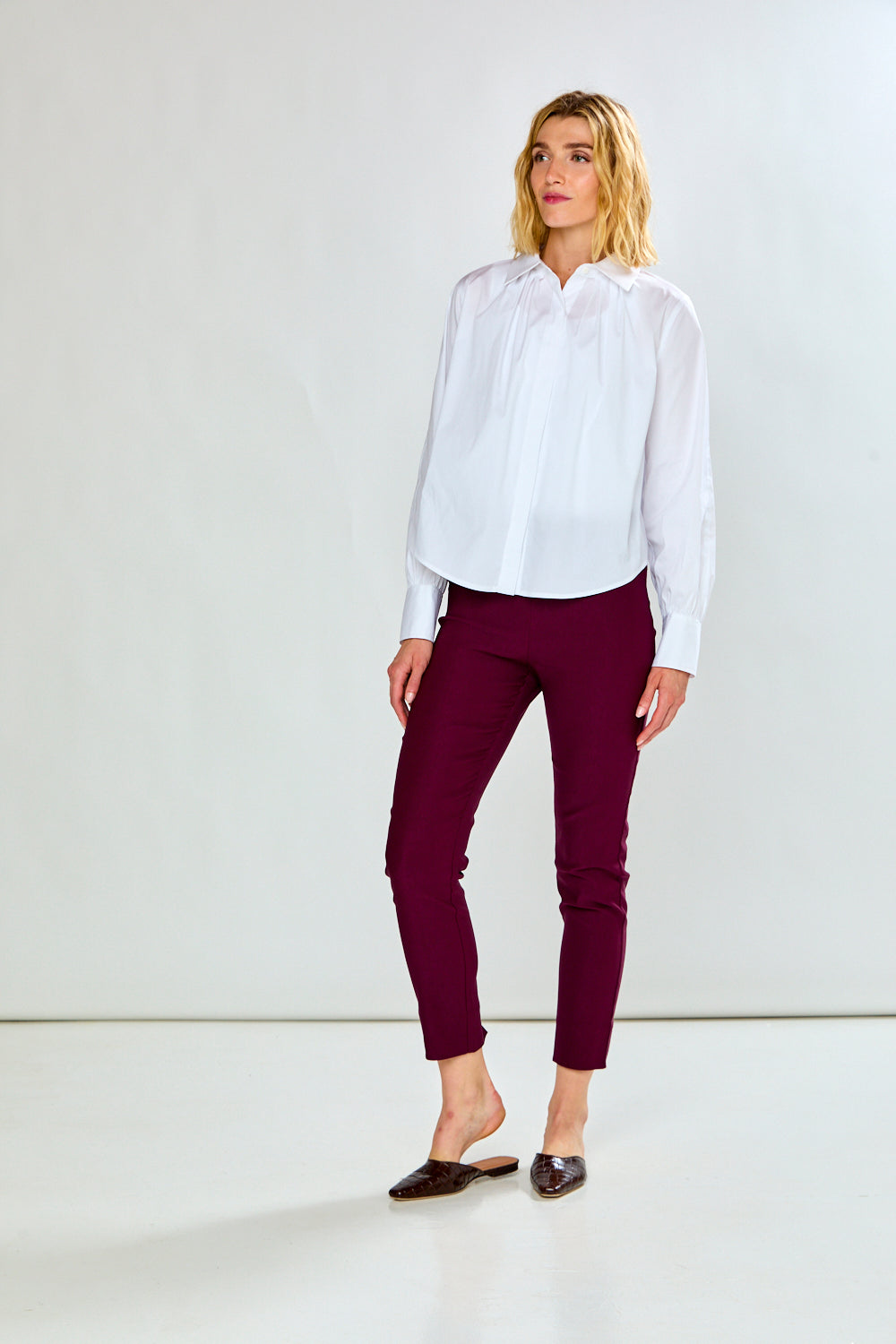A woman modeling a white long-sleeve button-up blouse and dark burgundy cropped pants, paired with dark brown mule flats, against a plain white studio background.