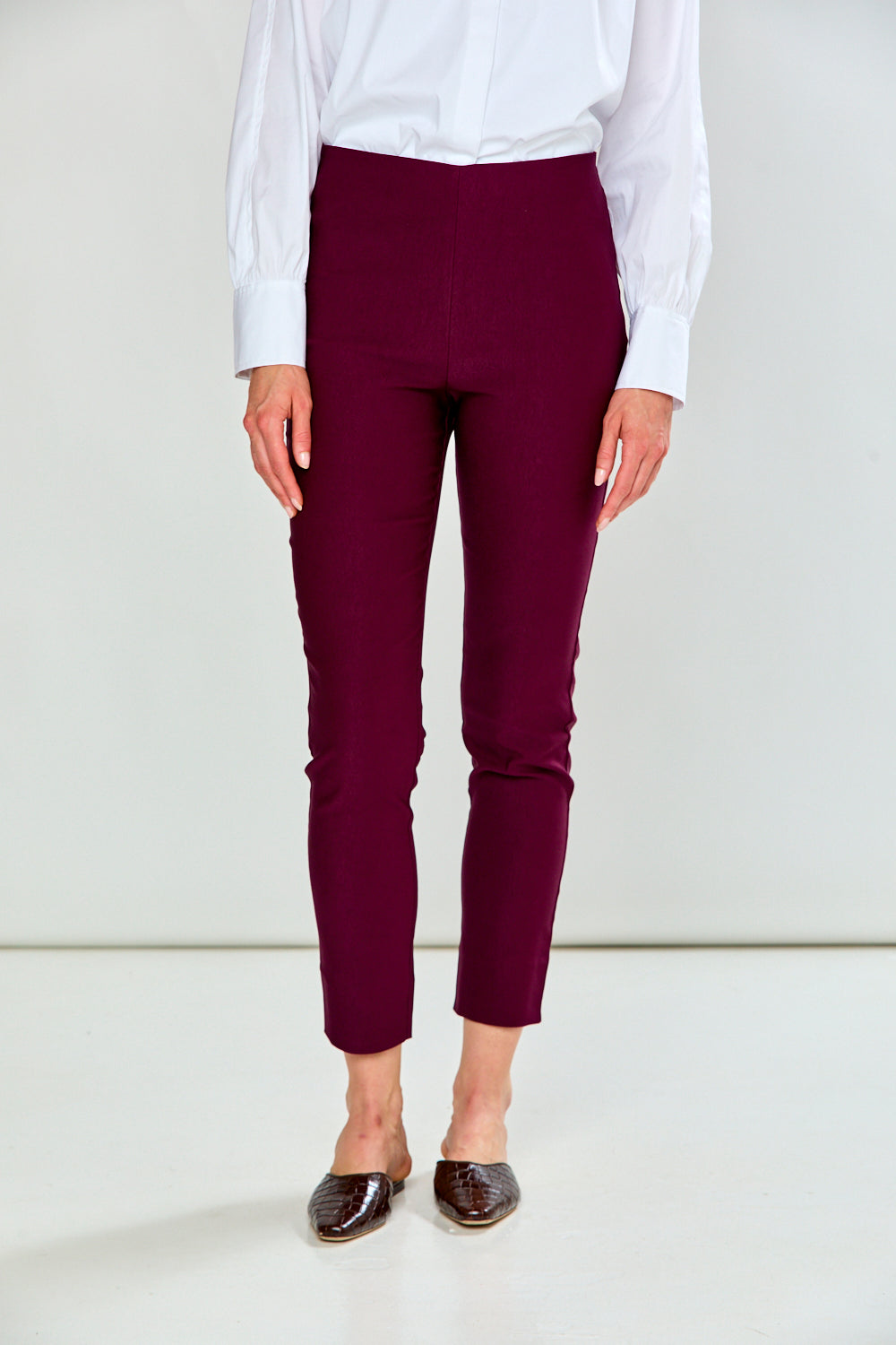 A model wearing high-waisted, cropped burgundy pants, paired with a white long-sleeve blouse and brown pointed-toe flats, shown from the waist down against a plain white studio background.